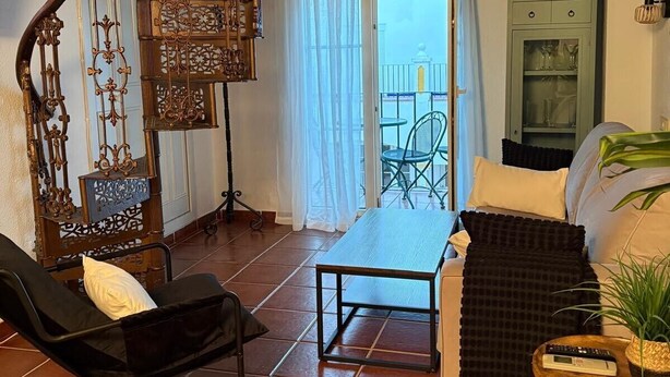 Ein Wohnzimmer mit einem schwarzen Loungesessel, einem Holztisch, einer Couch und einer Wendeltreppe. Es gibt einen Balkon mit Tisch und Stühlen sowie einen Schrank mit Glastüren.