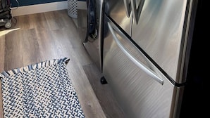 A compact kitchen with a stainless steel refrigerator, a built-in washer and dryer, a wicker basket, and a window with blinds.