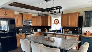 A modern kitchen with wooden cabinets, a central island with bar stools, a stainless steel refrigerator, and a hanging light fixture.