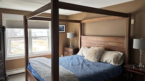 A four-poster bed with a headboard, a bedside table with a lamp, and a window with blinds.