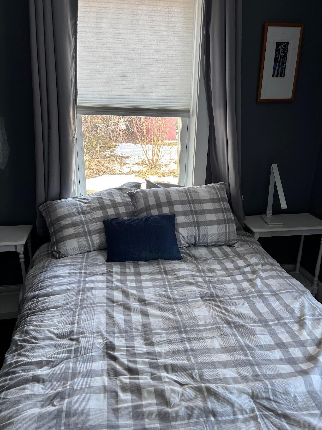 Una cama con colcha a cuadros, dos almohadas, una ventana con cortinas, un cuadro en la pared y una mesita de noche.