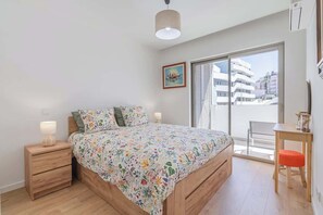 Un dormitorio con cama, mesitas de noche, una silla, ventana con persianas y vista a edificios.