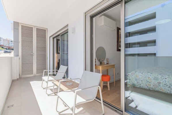 A balcony with a table and chairs, a bed with a floral bedspread, and a view of a building across the street.