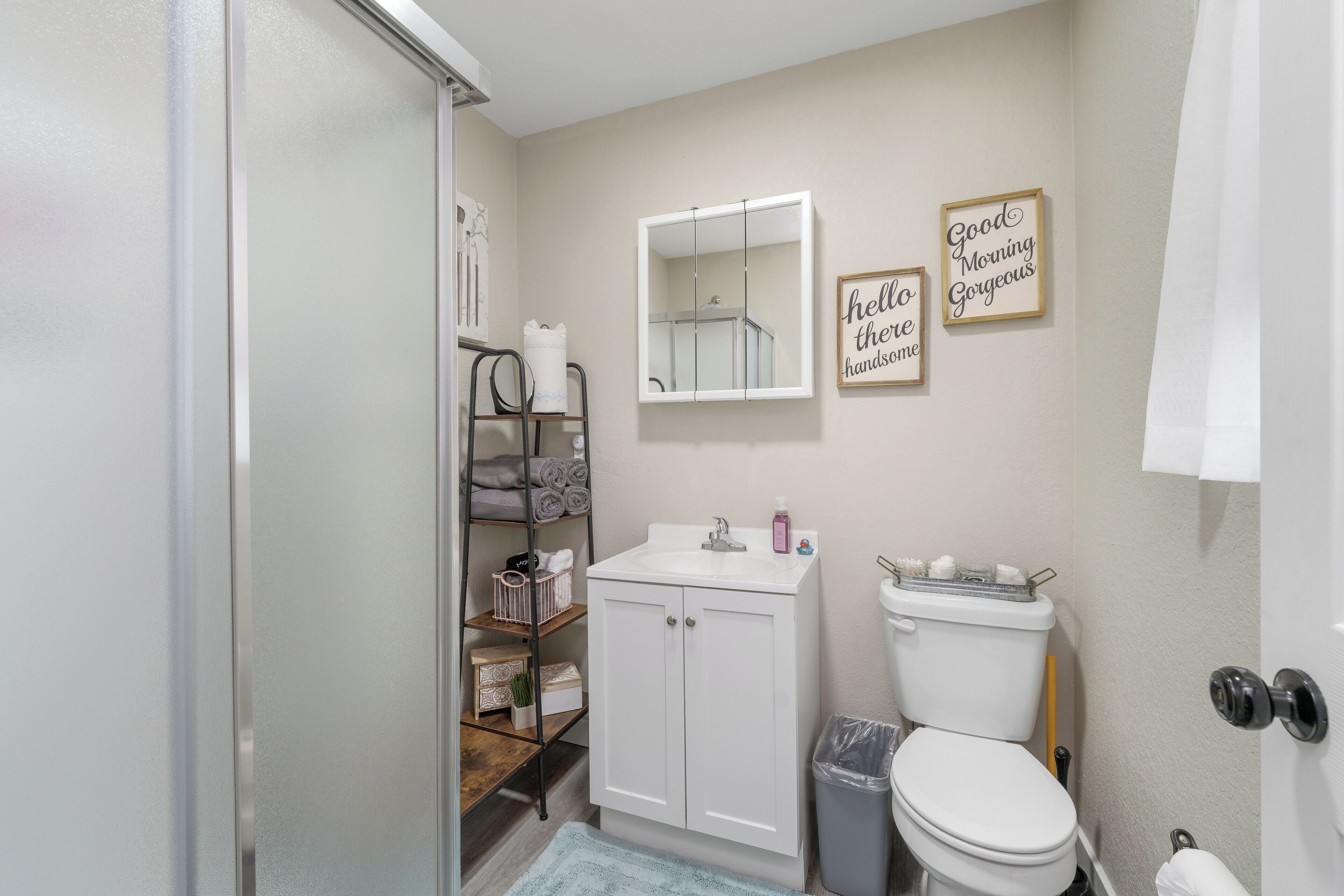 A bathroom with a white sink, a mirror, a toilet, a shelf with towels, and two motivational signs on the wall.