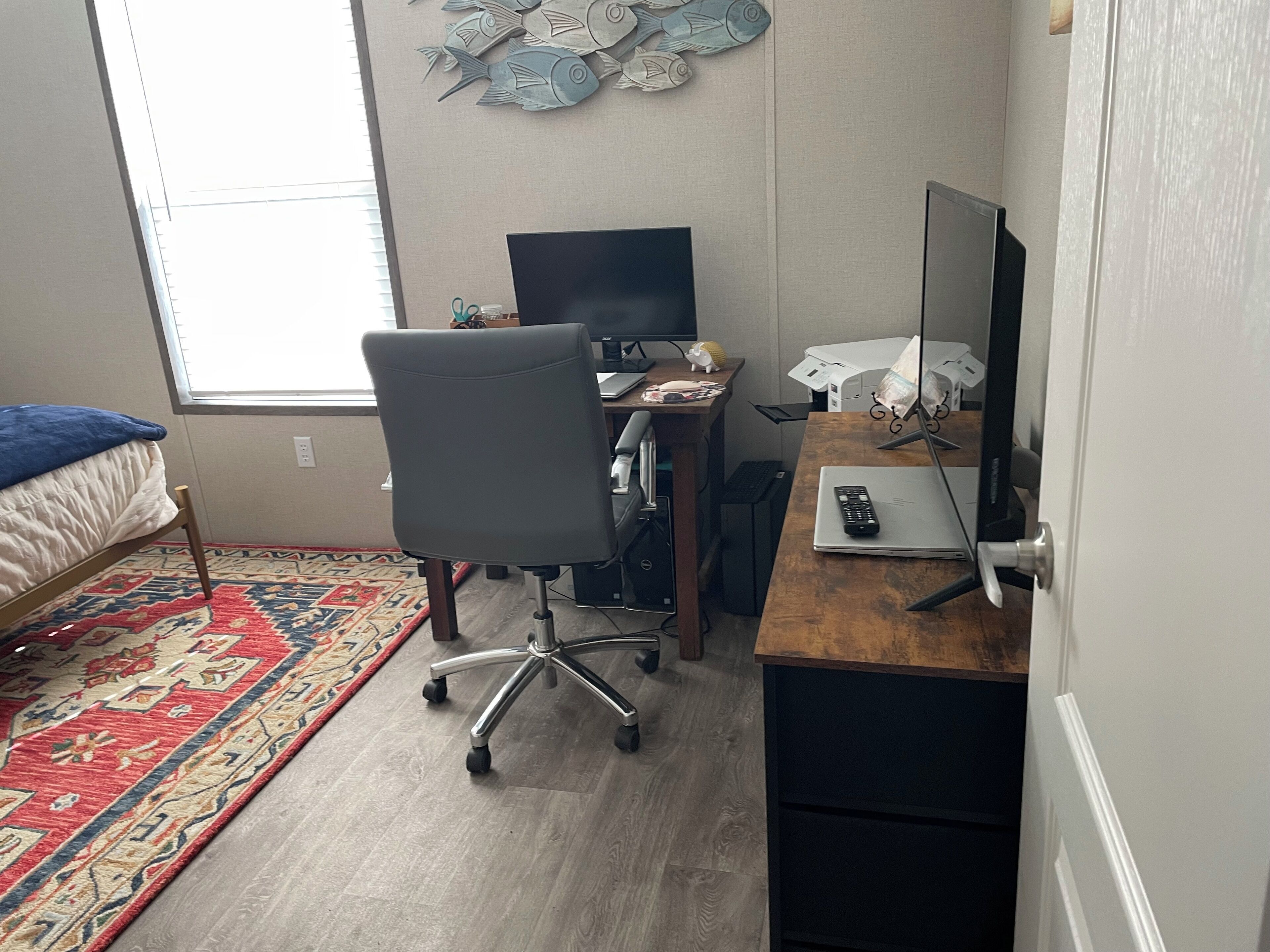 A small, well-lit room with a bed, a desk with a computer, and a chair. There is a window providing natural light and a decorative fish wall hanging.