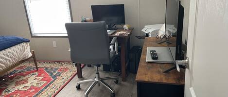A small, well-lit room with a bed, a desk with a computer, and a chair. There is a window providing natural light and a decorative fish wall hanging.