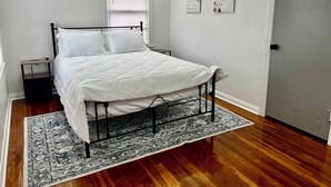 Uma cama bem arrumada com roupa de cama branca, estrutura de metal preto e um tapete estampado sobre piso de madeira.