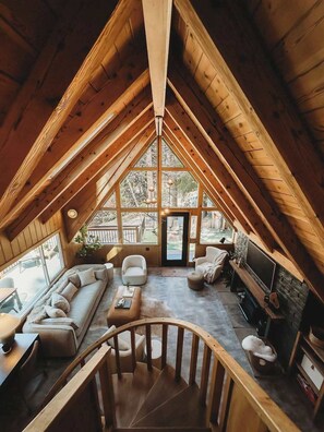 A cozy attic room with a wooden ceiling, a large window, a sofa, a TV, and a staircase.