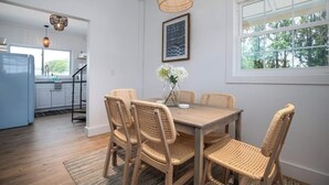 A dining area with a wooden table and wicker chairs, a vase with flowers, and a large window with a view of trees. - Ocean Boulevard South, 3 min walk to the beach! (Rye)