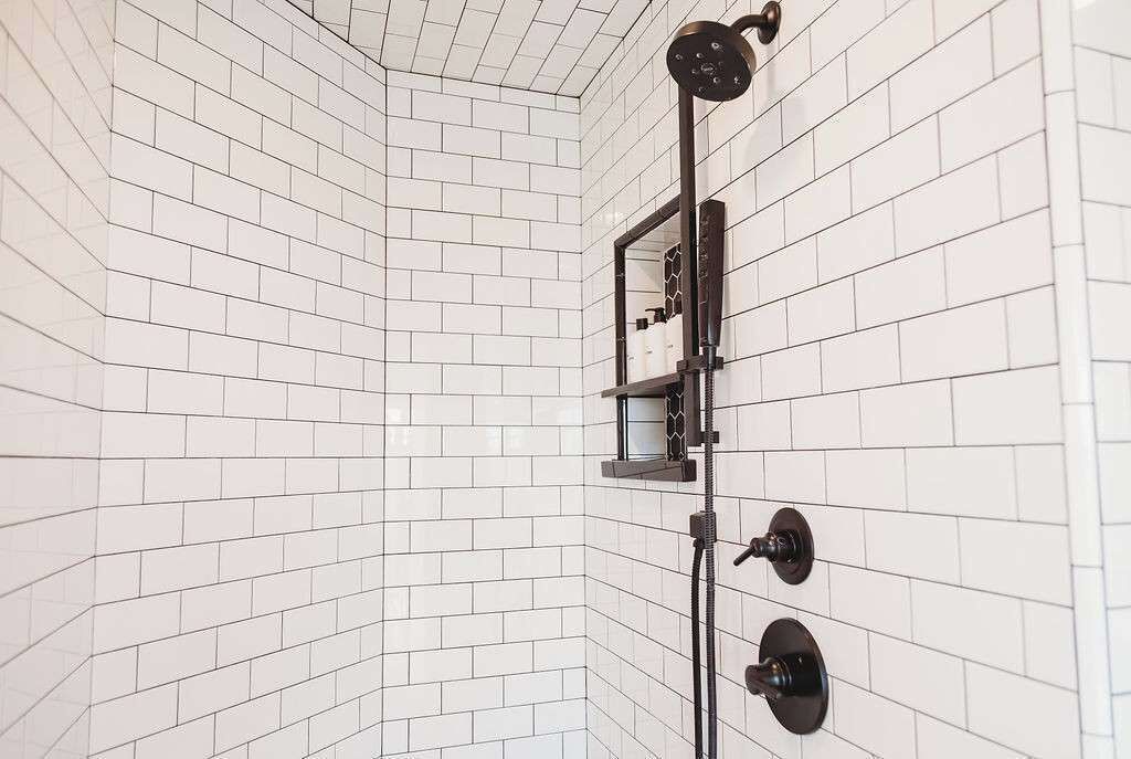 Eine Dusche mit schwarzem Duschschlauch und Armaturen, weißen Fliesenwänden und einem Eck-Duschkopf.