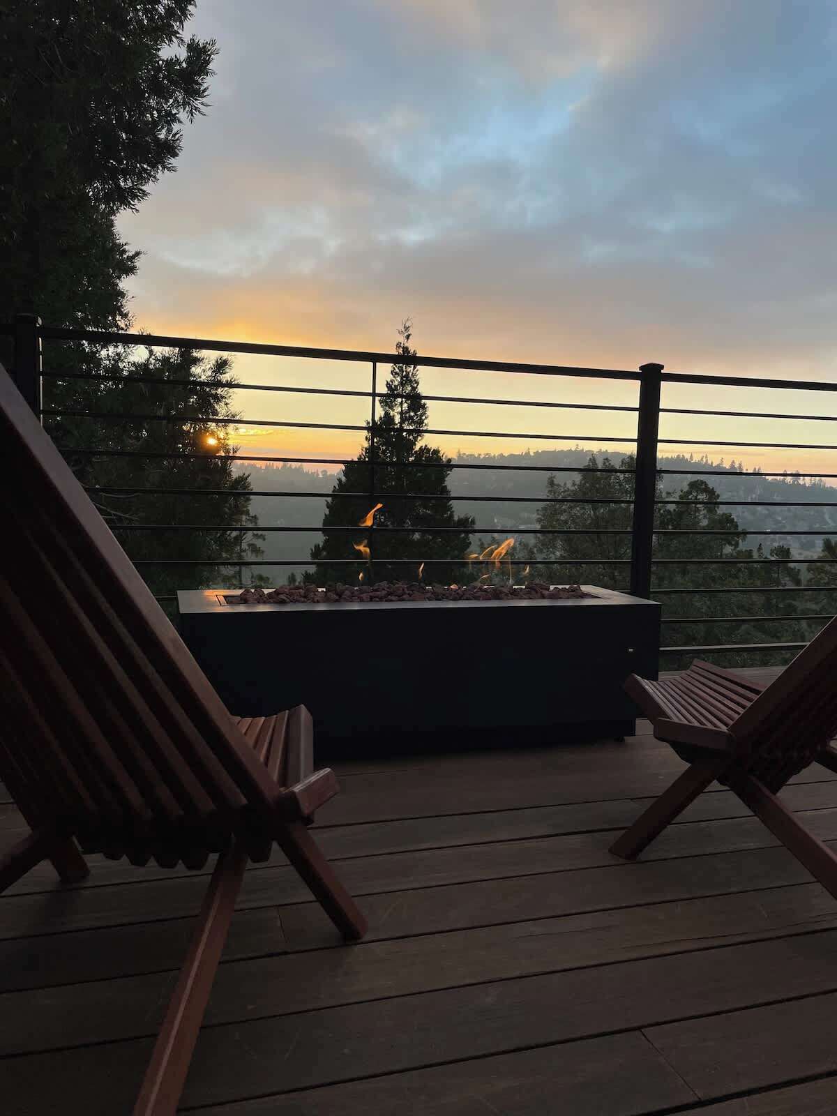 En terrasse med to træloungestole, en integreret pejs og udsigt til træer og en solnedgang.