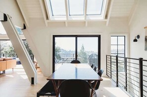 Um interior moderno com uma mesa de jantar, cadeiras, uma escada e janelas amplas que oferecem vista para o exterior.