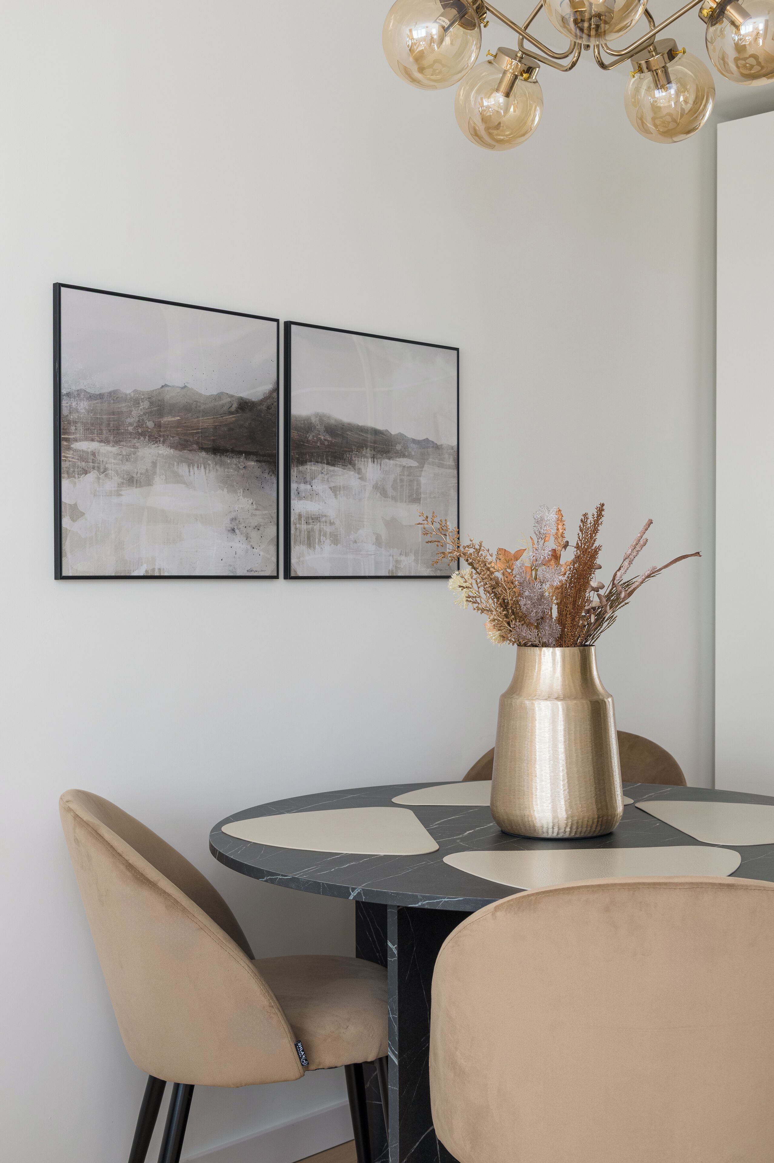 Un comedor con una mesa redonda, sillas color beige y un jarrón con flores secas. Dos cuadros están colgados en la pared.