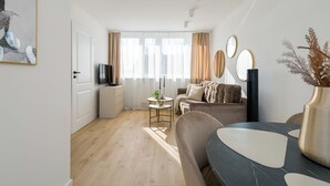 Een moderne woonkamer met een bank, een salontafel en een televisie. Er is een deur naar een keuken en een raam met gordijnen.