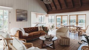 A spacious living room with a brown leather sofa, a white armchair, a round coffee table, and a dining area with a wooden table and chairs. - Palisades Point Lakehouse, by @To_Dwell_Here (Lake Arrowhead)