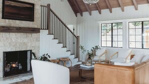 A spacious living room with a stone fireplace, a staircase, and a large window. - Palisades Point Lakehouse, by @To_Dwell_Here (Lake Arrowhead)