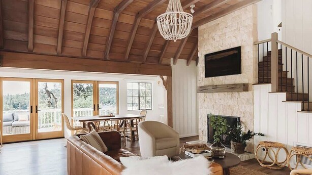 A spacious living room with a wooden ceiling, a fireplace, a television mounted on the wall, and a balcony with a view of the outdoors. - Palisades Point Lakehouse, by @To_Dwell_Here (Lake Arrowhead)
