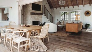 A spacious living area with a wooden dining table, wicker chairs, a sofa, and a staircase leading to an upper level. - Palisades Point Lakehouse, by @To_Dwell_Here (Lake Arrowhead)
