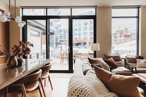 A modern hotel room with a large sliding glass door leading to an outdoor terrace, a sofa set, a dining table, and a vase with dried flowers.