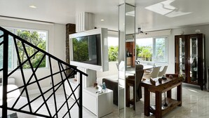 Uma sala de estar moderna com uma televisão de tela plana grande, um espaço de refeições com mesa de madeira e uma escada com corrimão geométrico.