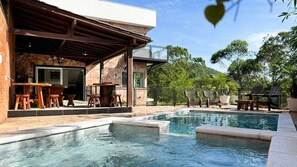 Zona de piscina con muro de piedra, mobiliario de madera y vistas a árboles y colinas.
