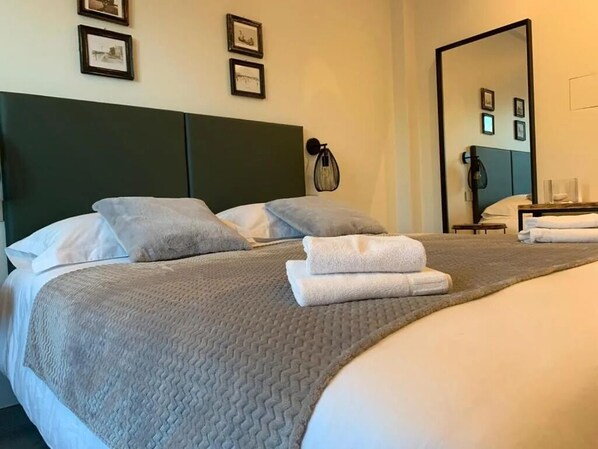 Una cama bien hecha con colcha gris y almohadas blancas, una mesita de noche con lámpara y cuadros en la pared.