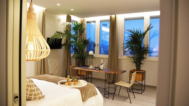 Ein modernes Hotelzimmer mit Bett, Frühstückswagen, Esstisch, Stühlen, Topfpflanzen und großen Fenstern.