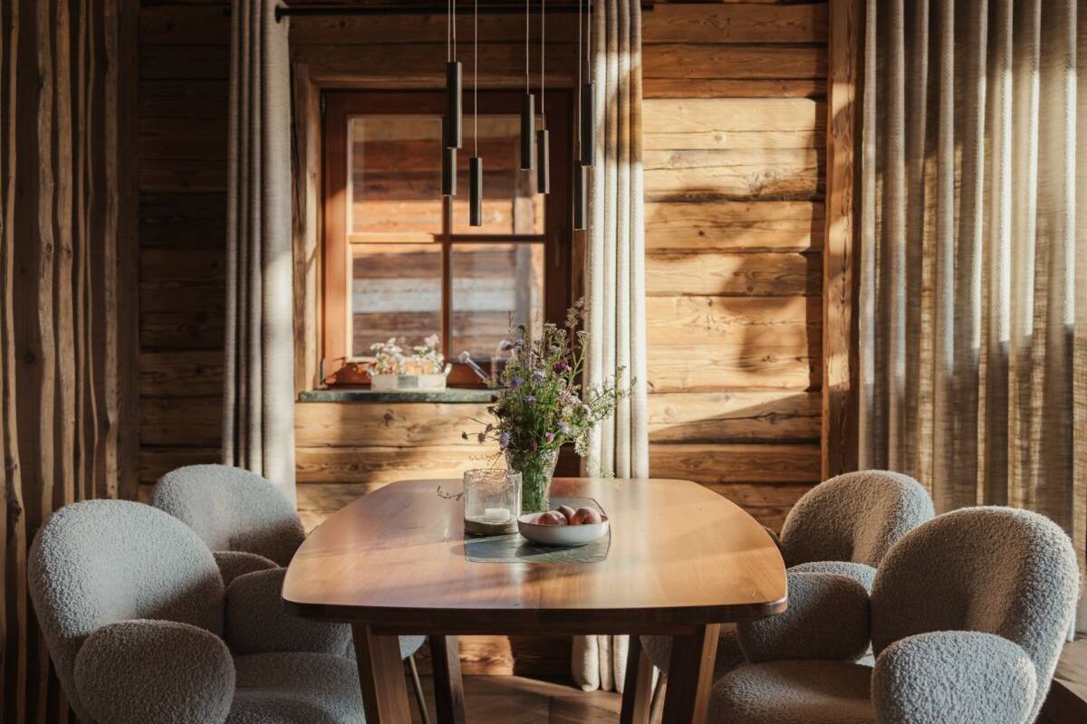 A wooden interior with a round table, grey upholstered chairs, a vase with flowers, and a window with curtains.
