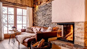 Un salon chaleureux avec un mur en pierre, une cheminée, un canapé en cuir et une fenêtre donnant sur des arbres couverts de neige.