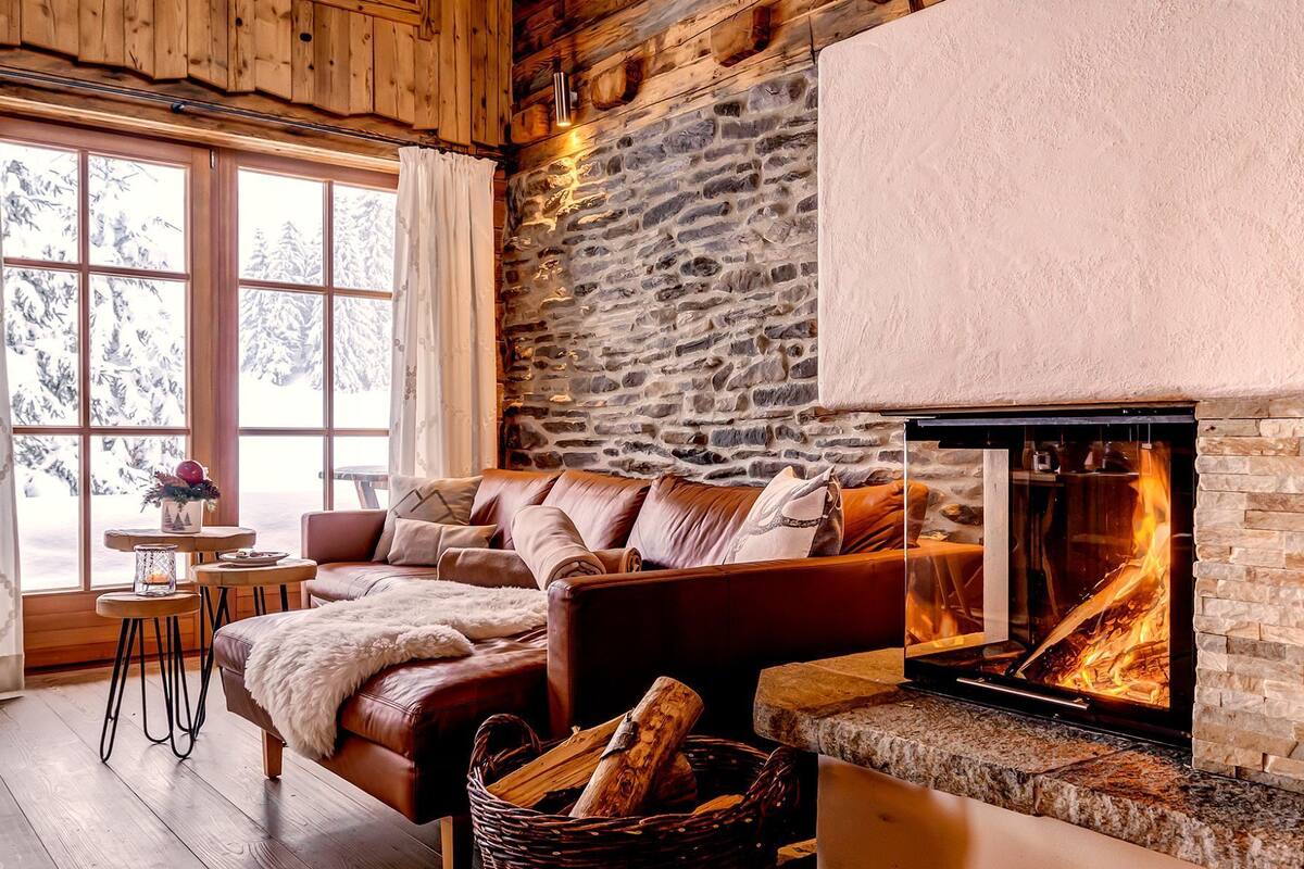 A cozy living room with a stone wall, a fireplace, a leather sofa, and a window with a view of snow-covered trees.