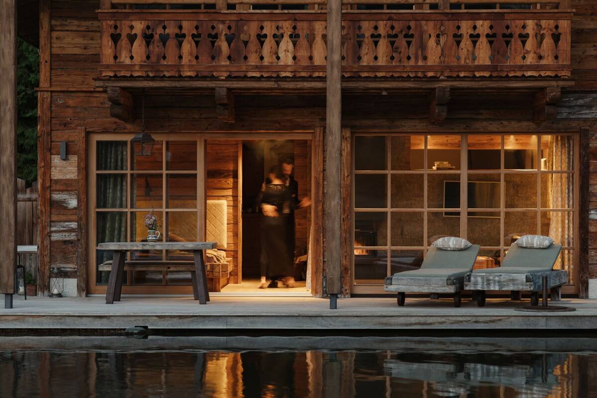 A wooden cabin with a balcony, a pool, and a lounge area with two chairs.