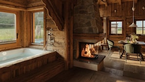 L’intérieur d’une cabane en rondins, avec une cheminée en pierre, une grande baignoire en bois et un coin repas offrant une vue sur l’extérieur.