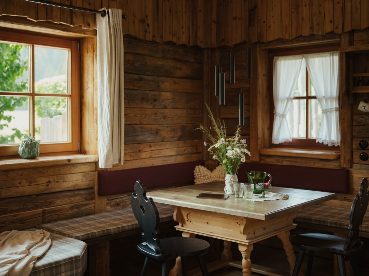 A wooden cabin interior with a wooden table, chairs, a bench, and two windows with curtains.