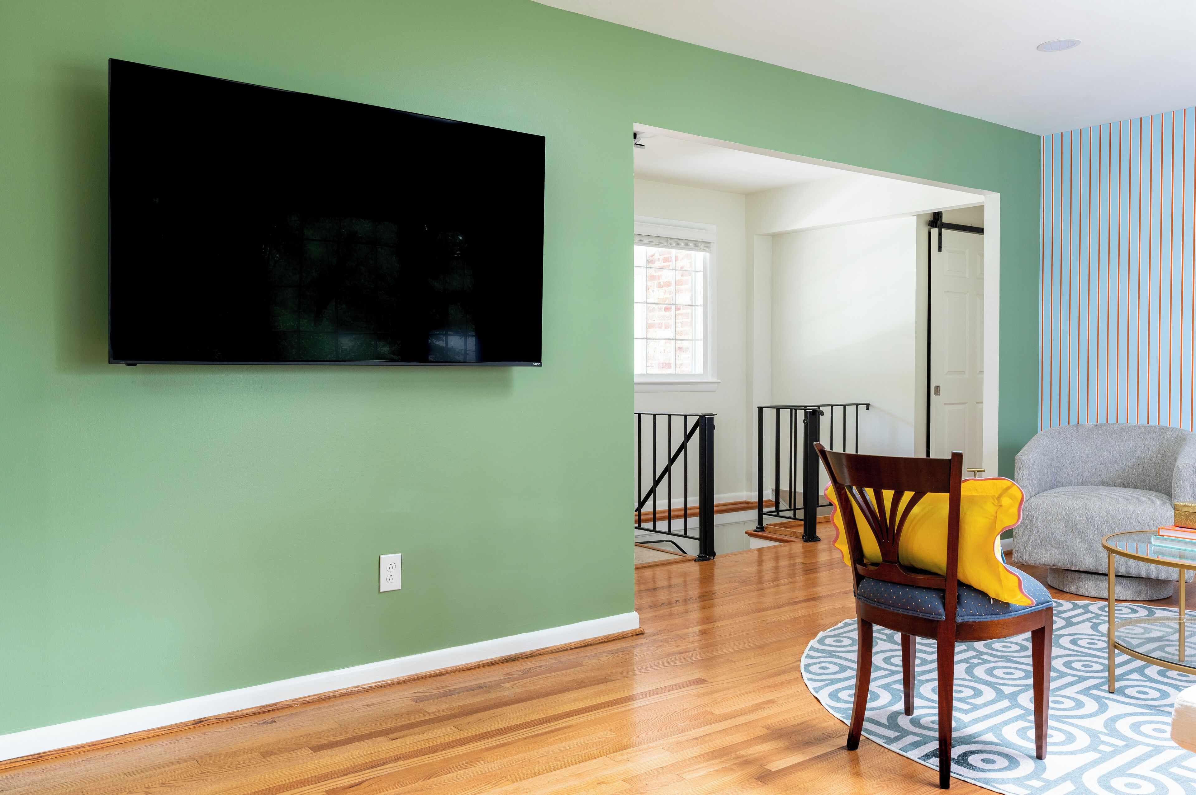 Un salon avec un téléviseur à écran plat fixé sur un mur vert, une chaise en bois avec un coussin jaune, un canapé gris et un tapis à motifs.