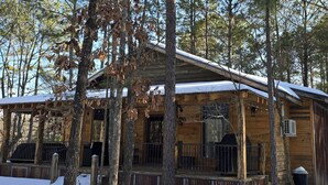 A wooden cabin with a porch, surrounded by trees and covered in snow. - Rustic Comfort Cabins-Anglers Basin with Hot Tub (Amity)