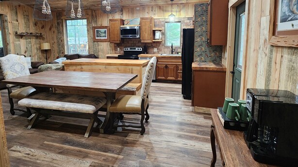 A cozy cabin-style interior with a wooden dining table, bench seating, a kitchen area with a microwave and stove, and a coffee maker on a side table. - Rustic Comfort Cabins-Anglers Basin with Hot Tub (Amity)