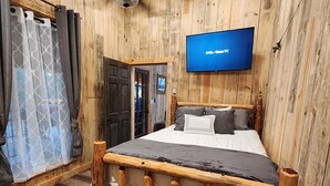 A wooden-paneled room with a bed, a TV mounted on the wall, and a wooden bench. - Rustic Comfort Cabins-Anglers Basin with Hot Tub (Amity)