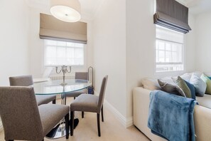 Un salon moderne avec une table à manger au plateau en verre, des chaises à revêtement gris, un canapé beige agrémenté d’un plaid bleu et une fenêtre ornée d’un lambrequin foncé.