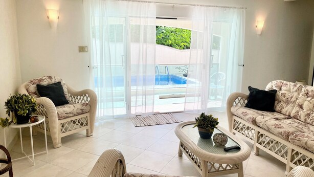 Ein geräumiges Wohnzimmer mit zwei Rattan-Sofas, einem Couchtisch und einer Topfpflanze. Eine Glastür öffnet sich zu einem Poolbereich.