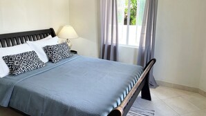 Ein ordentlich gemachtes Bett mit blauer Bettdecke und gemusterten Kissen, eine Nachttischlampe und ein Fenster mit Vorhängen.