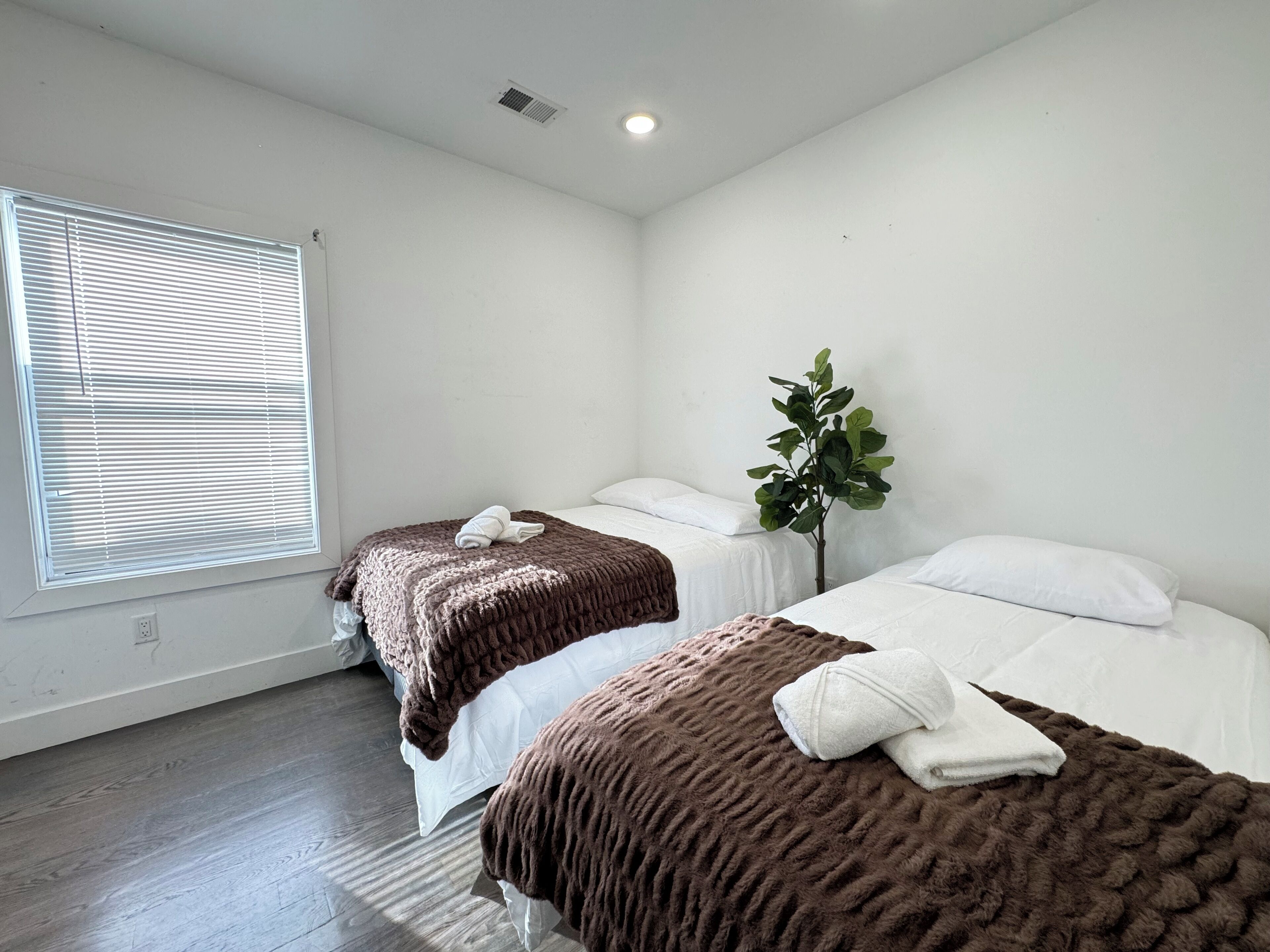 A hotel room with two beds, a window with blinds, a plant, and a ceiling vent.