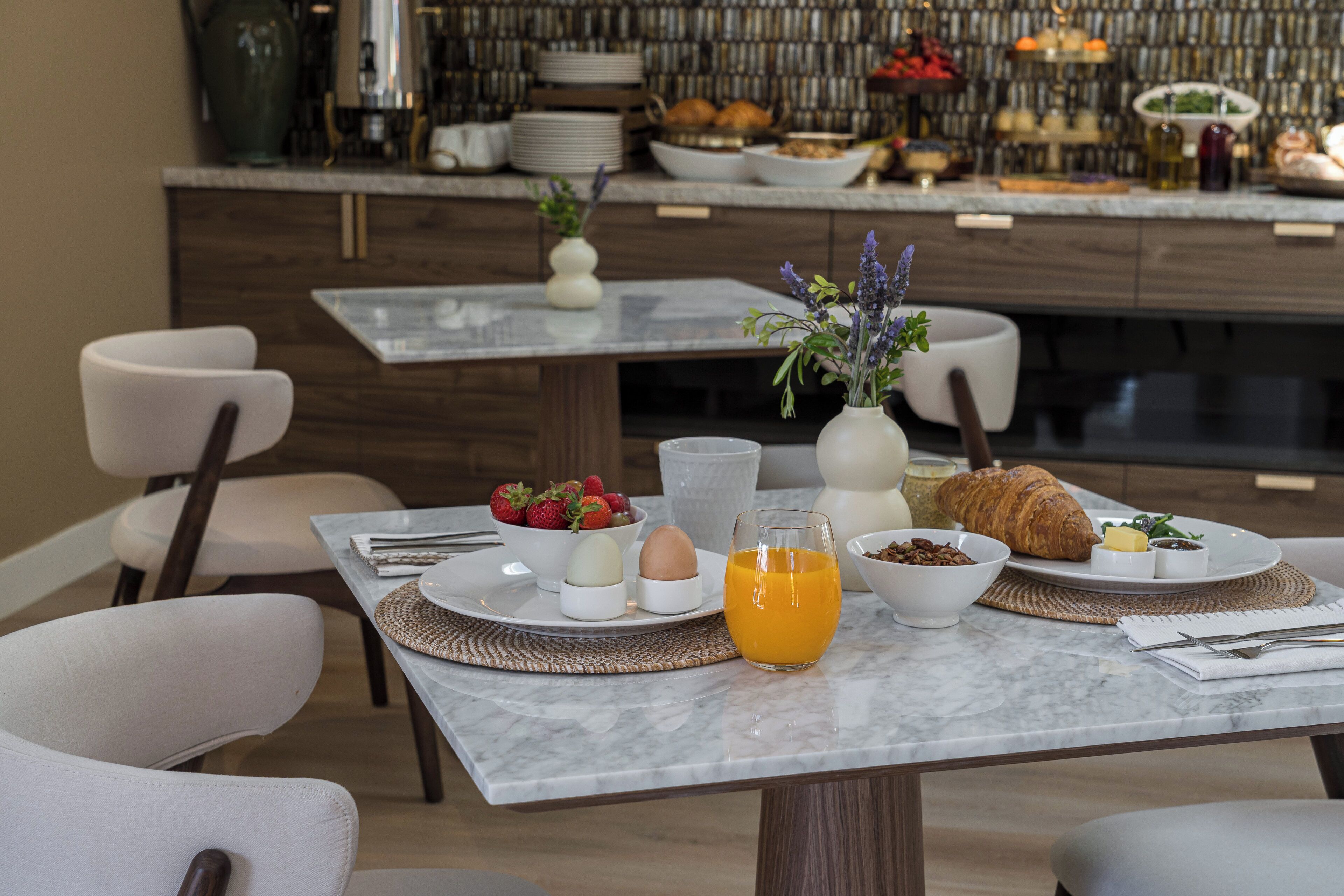 A breakfast setting with a marble table, white chairs, and a variety of breakfast items including eggs, fruit, and pastries.