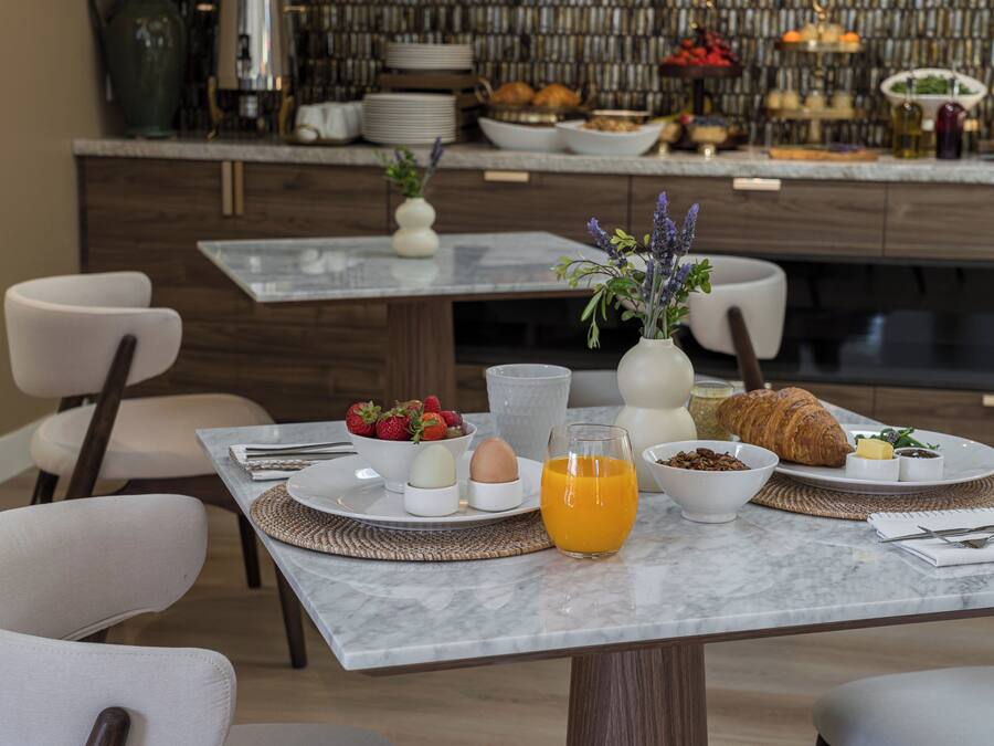 A breakfast setting with a marble table, white chairs, and a variety of breakfast items including eggs, fruit, and pastries.