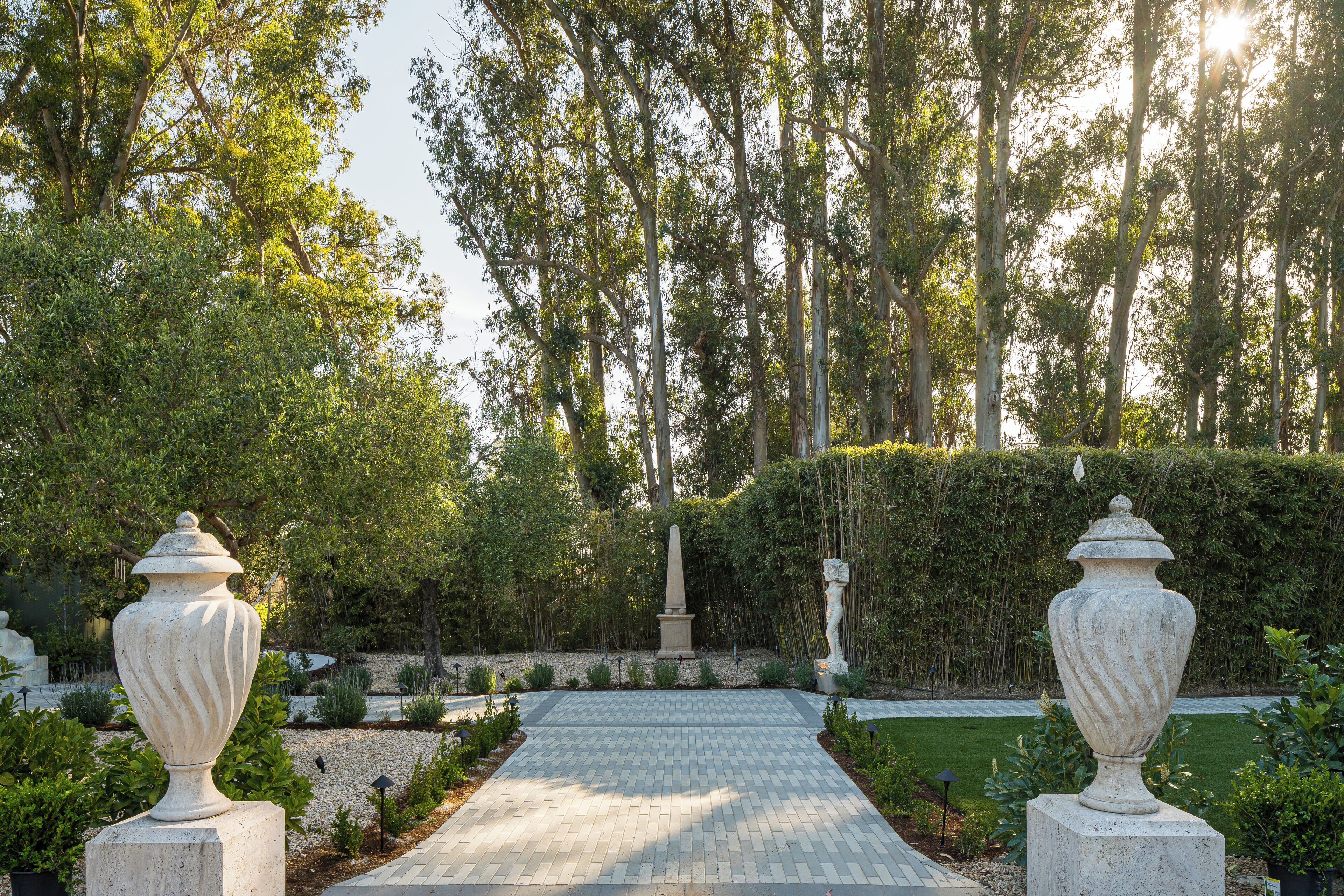 A paved pathway leading through a garden with two large urns on either side, surrounded by greenery and a hedge.