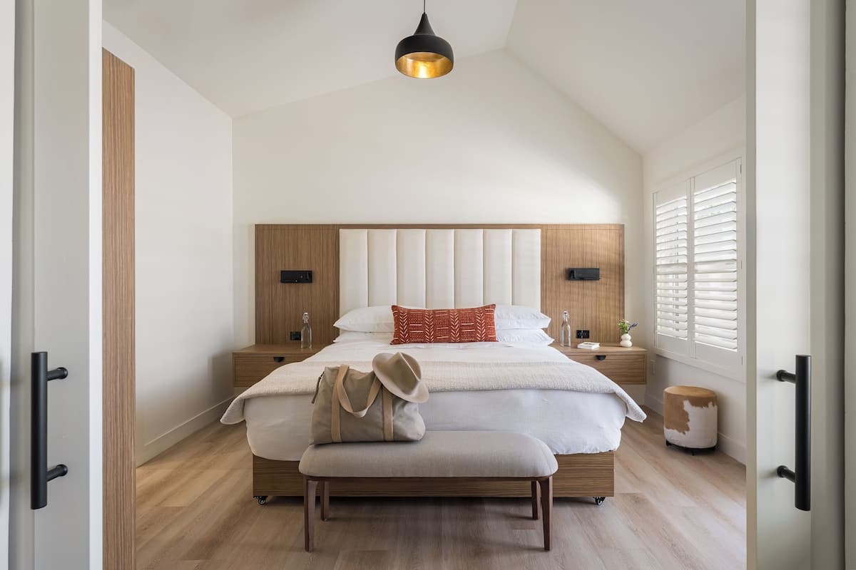 A bedroom with a large bed, a bench, a hanging light fixture, and a window with blinds.