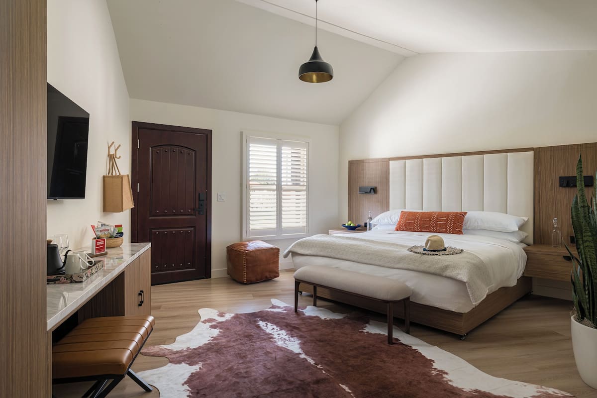 A bedroom with a large bed, a desk, a chair, a cowhide rug, a wooden door, and a ceiling light.