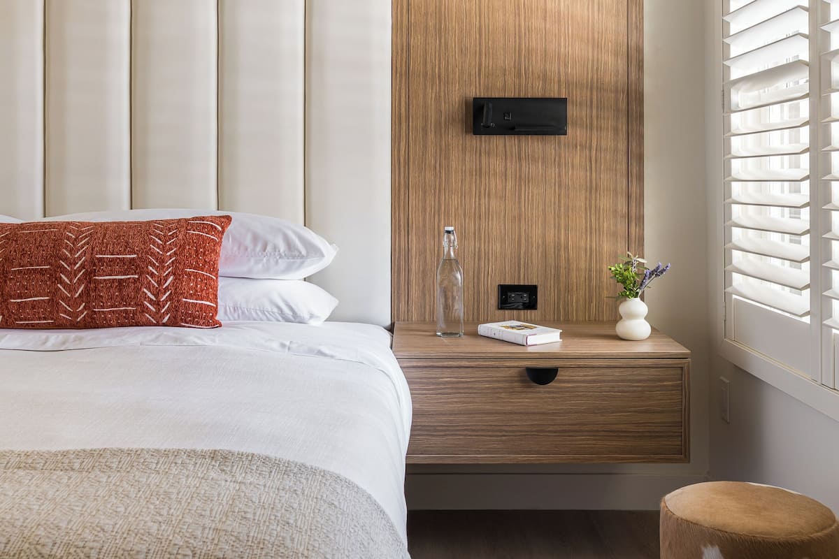 A modern bedroom with a large bed, a wooden headboard, a bedside table with a bottle, a book, and a vase.