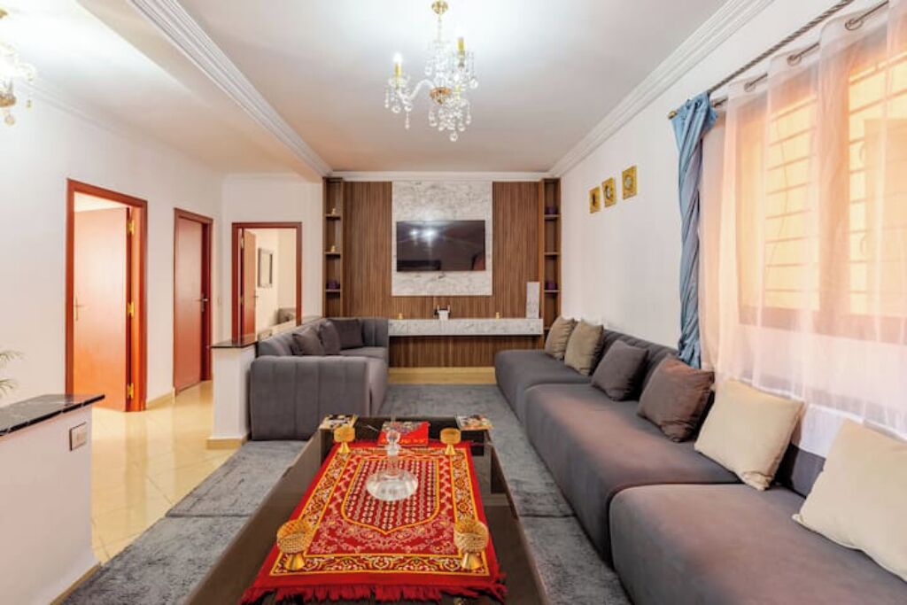 A living room with a grey sofa, a coffee table with a decorative rug, a chandelier, and a TV mounted on the wall.