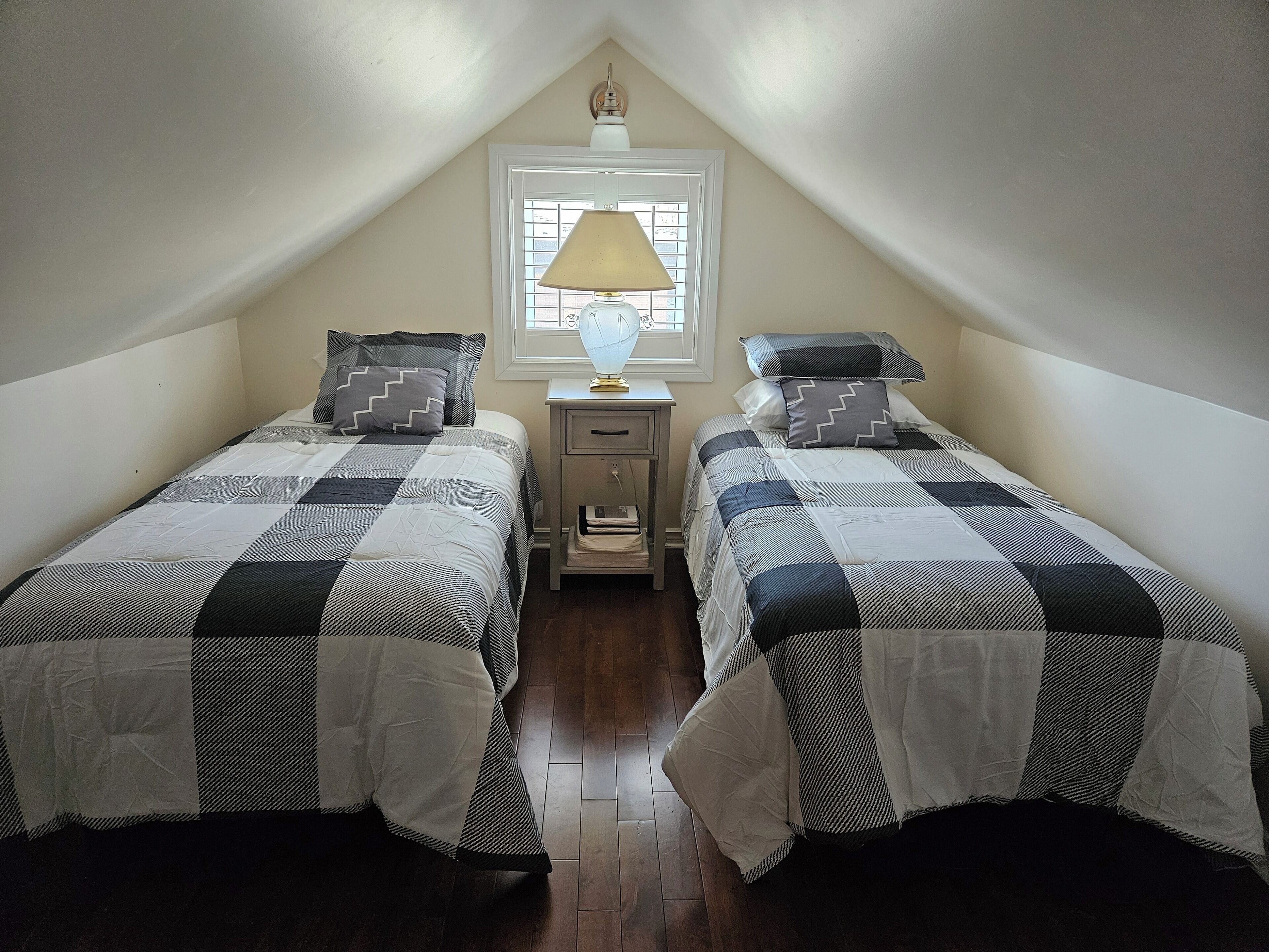 A small, cozy attic bedroom with two beds, a nightstand, a lamp, and a window.