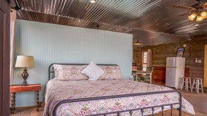 A bedroom with a metal bed frame, a wooden nightstand with a lamp, a floral bedspread, and a wooden ceiling with exposed beams. - Leaning Tower of Alpine (Alpine)